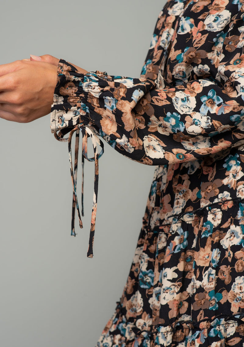 [Color: Black/Natural] A close up image of a brunette model wearing a bohemian mini dress in a black and natural floral print. With long sleeves, adjustable wrist cuffs, and a flowy tiered mini skirt. 