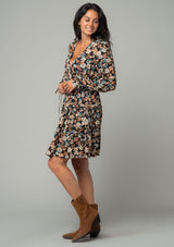 [Color: Black/Natural] A side facing image of a brunette model wearing a bohemian mini dress in a black and natural floral print. With long sleeves, adjustable wrist cuffs, and a flowy tiered mini skirt. 