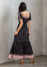 [Color: Black/Rose] A model wearing a black bohemian maxi dress with a pink floral border print. With short puff sleeves, a square neckline, and a tiered skirt. A romantic Spring dress.