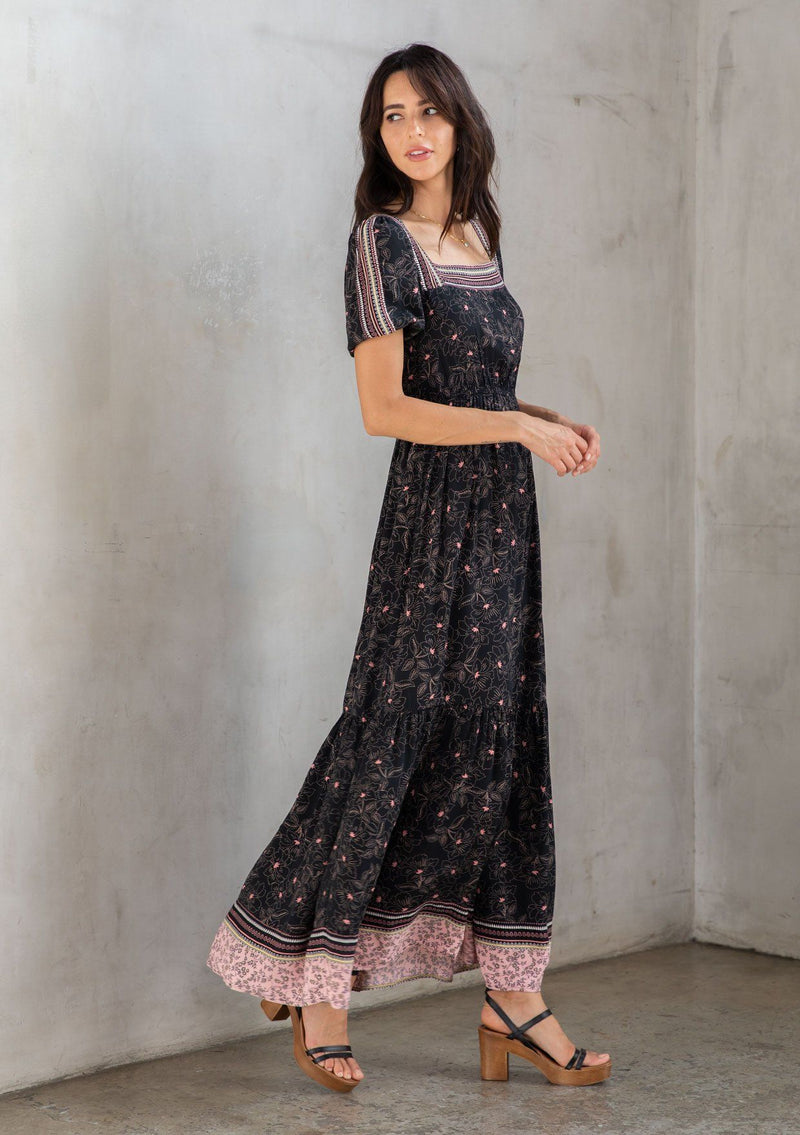 [Color: Black/Rose] A model wearing a black bohemian maxi dress with a pink floral border print. With short puff sleeves, a square neckline, and a tiered skirt. A romantic Spring dress.