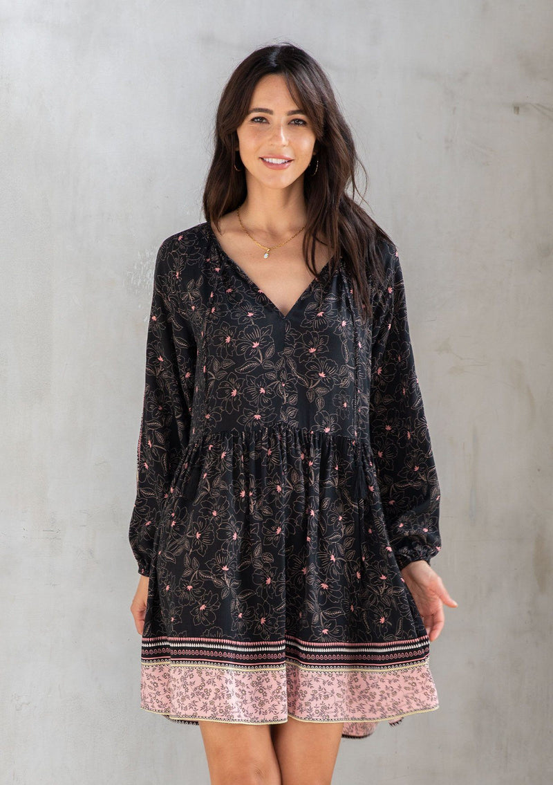 [Color: Black/Rose] A model wearing a classic black bohemian mini dress in a pink floral border print. With a loose, relaxed silhouette, long sleeves, and a split v neckline with tassels.