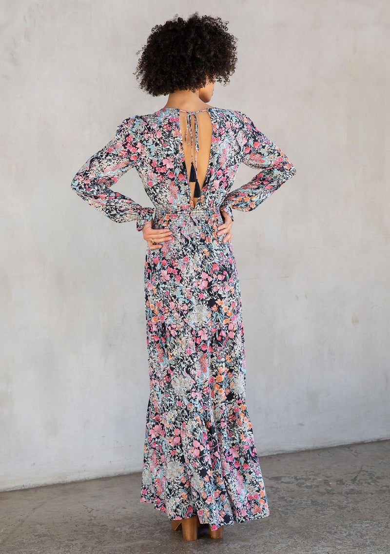 [Color: Black/Pink] A model wearing a timeless bohemian black and pink floral print maxi dress. With a deep v neckline in front and back, a tassel tie closure, and long sleeves. 