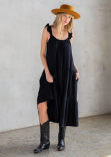[Color: Black] A model wearing a classic black maxi dress designed in soft cotton gauze. With a scooped neckline, adjustable ruffled straps, and side pockets.