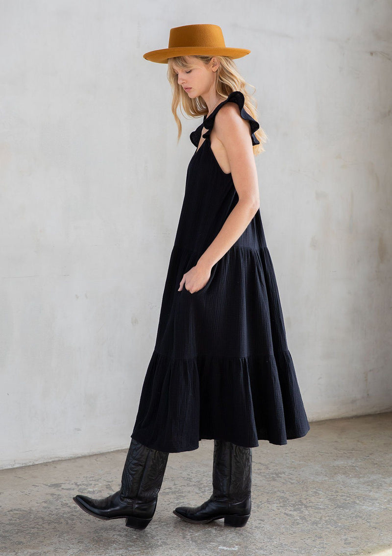 [Color: Black] A model wearing a classic black maxi dress designed in soft cotton gauze. With a scooped neckline, adjustable ruffled straps, and side pockets. 