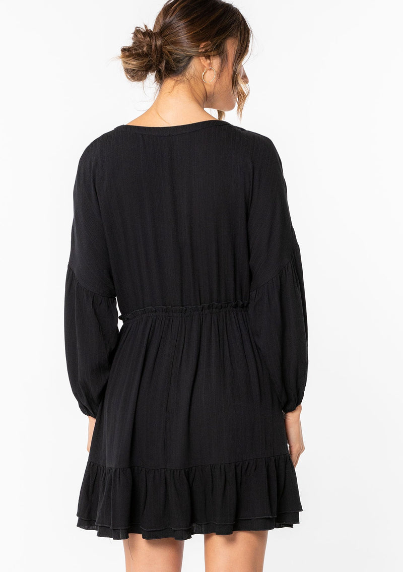 [Color: Black] A model wearing a simple black flowy bohemian mini dress with long sleeves and a button front. 