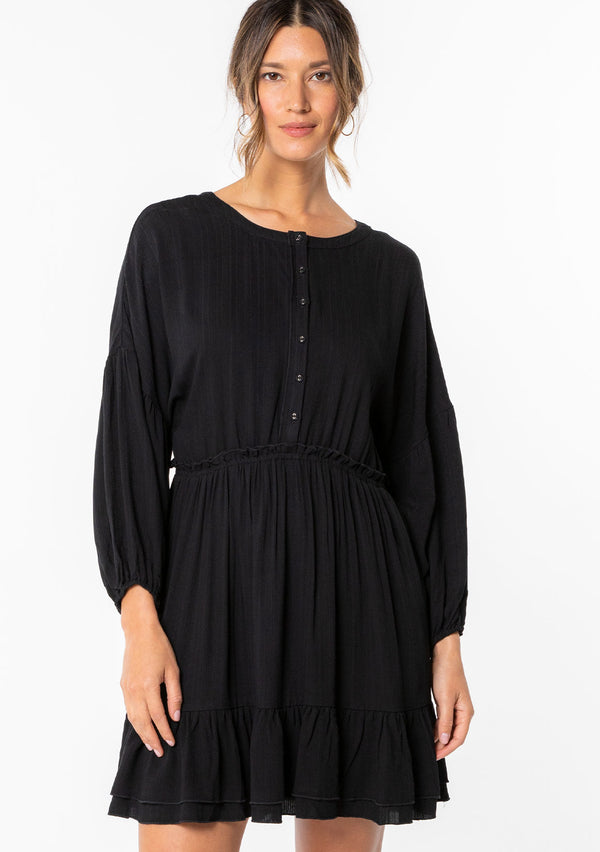 [Color: Black] A model wearing a simple black flowy bohemian mini dress with long sleeves and a button front. 