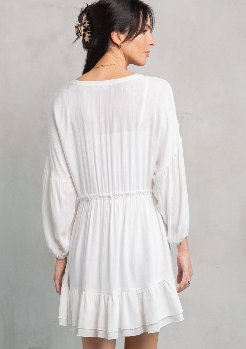 [Color: Off White] A model wearing a simple white flowy bohemian mini dress with long sleeves and a button front. 