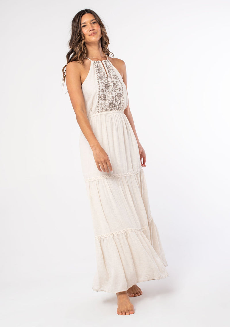 [Color: Off White/Taupe] A model wearing an off white flowy bohemian maxi dress with a halter tie neckline and embroidered front. 