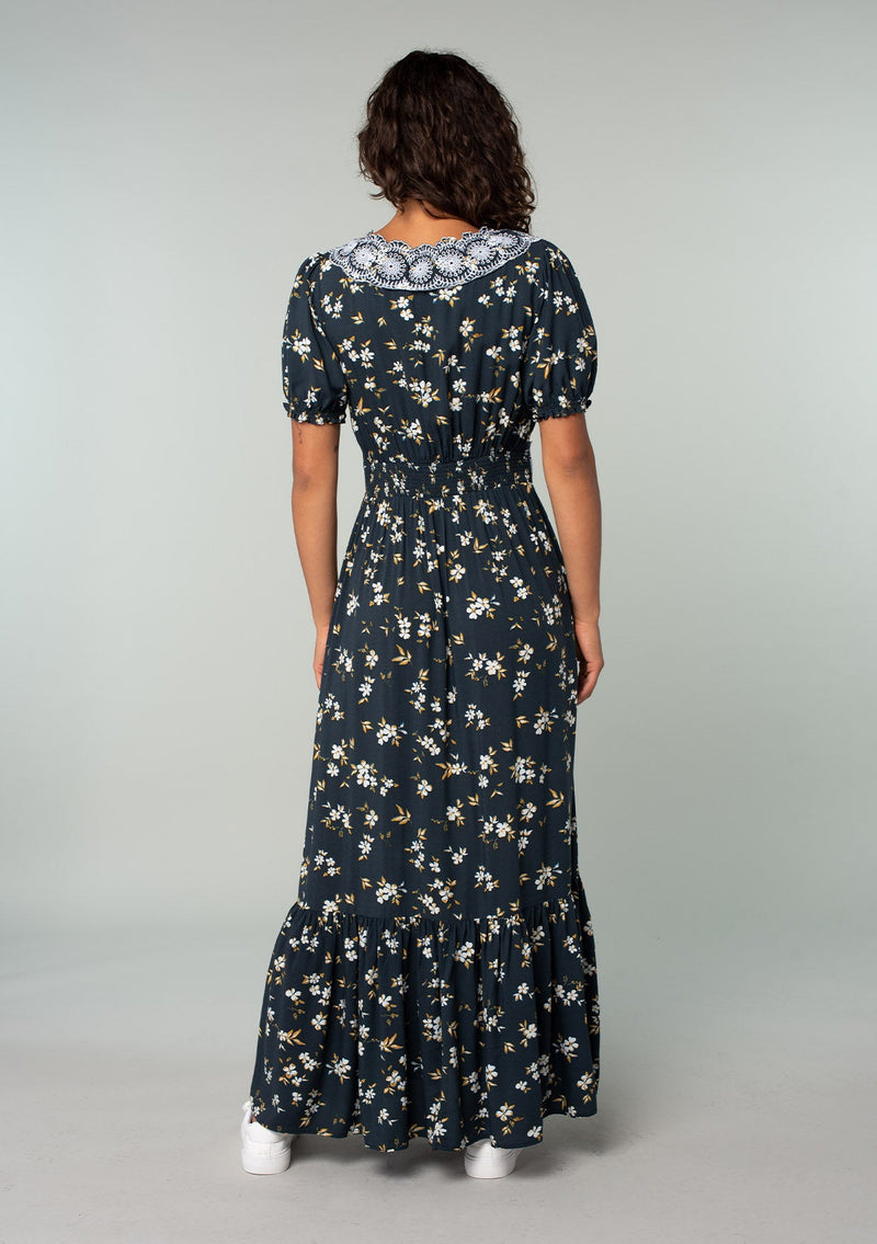 [Color: Navy/Powder Blue] A back facing image of a brunette model wearing a bohemian maxi dress in a navy blue and yellow floral print. With short puff sleeves, a lace trimmed surplice v neckline, a lace trimmed waist, and a button front skirt. 