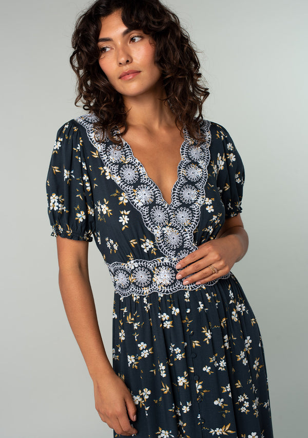 [Color: Navy/Powder Blue] A close up front facing image of a brunette model wearing a bohemian maxi dress in a navy blue and yellow floral print. With short puff sleeves, a lace trimmed surplice v neckline, a lace trimmed waist, and a button front skirt. 