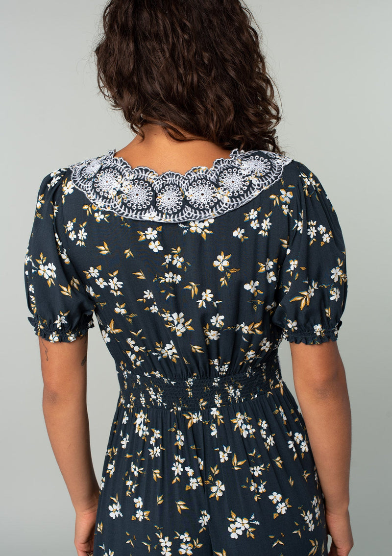 [Color: Navy/Powder Blue] A close up back facing image of a brunette model wearing a bohemian maxi dress in a navy blue and yellow floral print. With short puff sleeves, a lace trimmed surplice v neckline, a lace trimmed waist, and a button front skirt. 