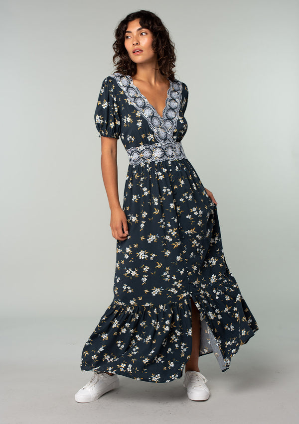 [Color: Navy/Powder Blue] A front facing image of a brunette model wearing a bohemian maxi dress in a navy blue and yellow floral print. With short puff sleeves, a lace trimmed surplice v neckline, a lace trimmed waist, and a button front skirt. 