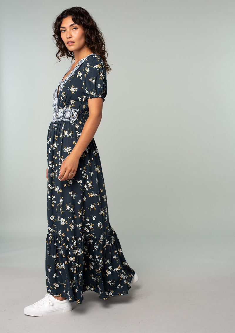 [Color: Navy/Powder Blue] A side facing image of a brunette model wearing a bohemian maxi dress in a navy blue and yellow floral print. With short puff sleeves, a lace trimmed surplice v neckline, a lace trimmed waist, and a button front skirt. 