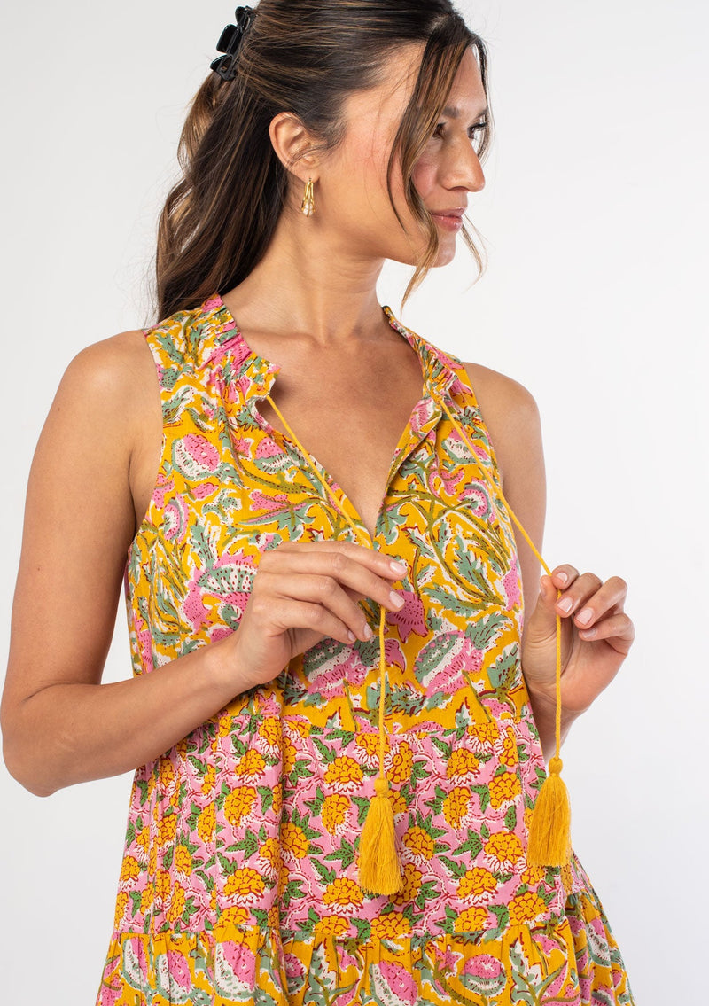 [Color: Pink/Mango] A woman wearing a bright pink and yellow floral print sleeveless tent dress with a tiered silhouette and tassel ties. 