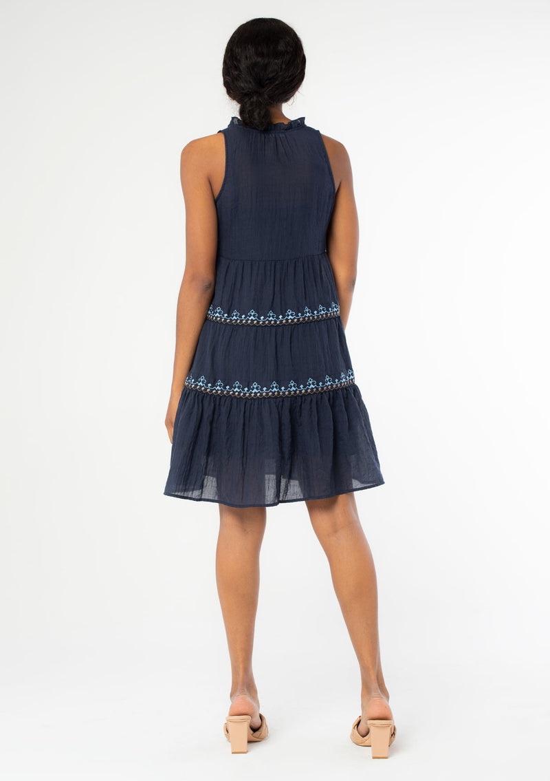 [Color: Navy] A back facing image of a black model wearing a navy blue cotton bohemian mini tent dress with tassel ties and light blue embroidered detail. 