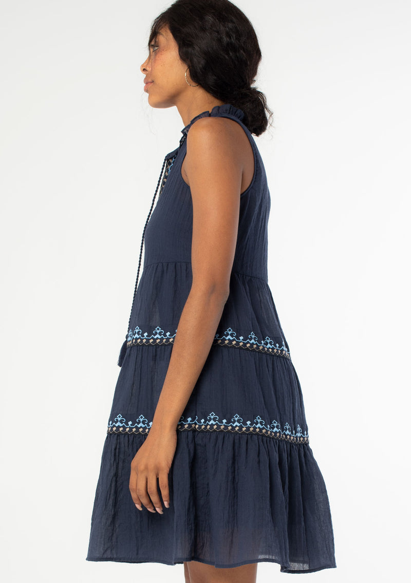 [Color: Navy] A side facing image of a black model wearing a navy blue cotton bohemian mini tent dress with tassel ties and light blue embroidered detail. 