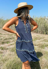 [Color: Navy] A front facing image of a brunette model outside wearing a navy blue cotton bohemian mini tent dress with tassel ties and light blue embroidered detail.