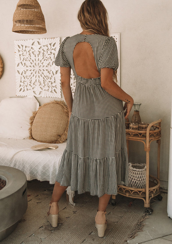[Color: Black/Natural] A model wearing a black and white small checkered gingham mid length dress with short puff sleeves and an open back detail with button closure.
