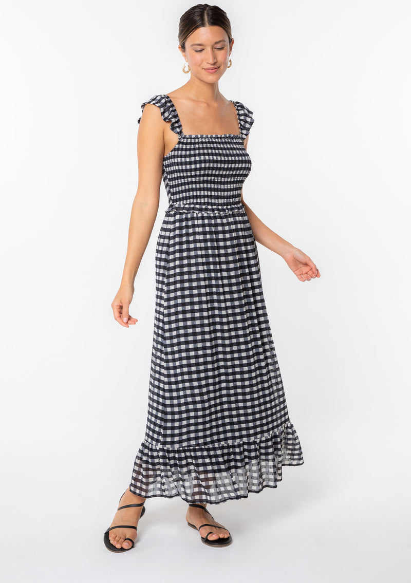 [Color: Black/Off White] A model wearing a black and white gingham checkered maxi dress in a clip dot chiffon. With ruffled tank top straps, a smocked bodice, and a square neckline. 