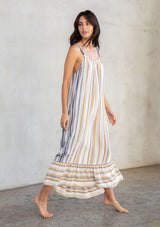 [Color: Dijon/Navy] A model wearing a vacation ready sleeveless maxi dress in a yellow and blue stripe. With a ruffled round neckline, a relaxed flowy fit, and a tiered skirt. 
