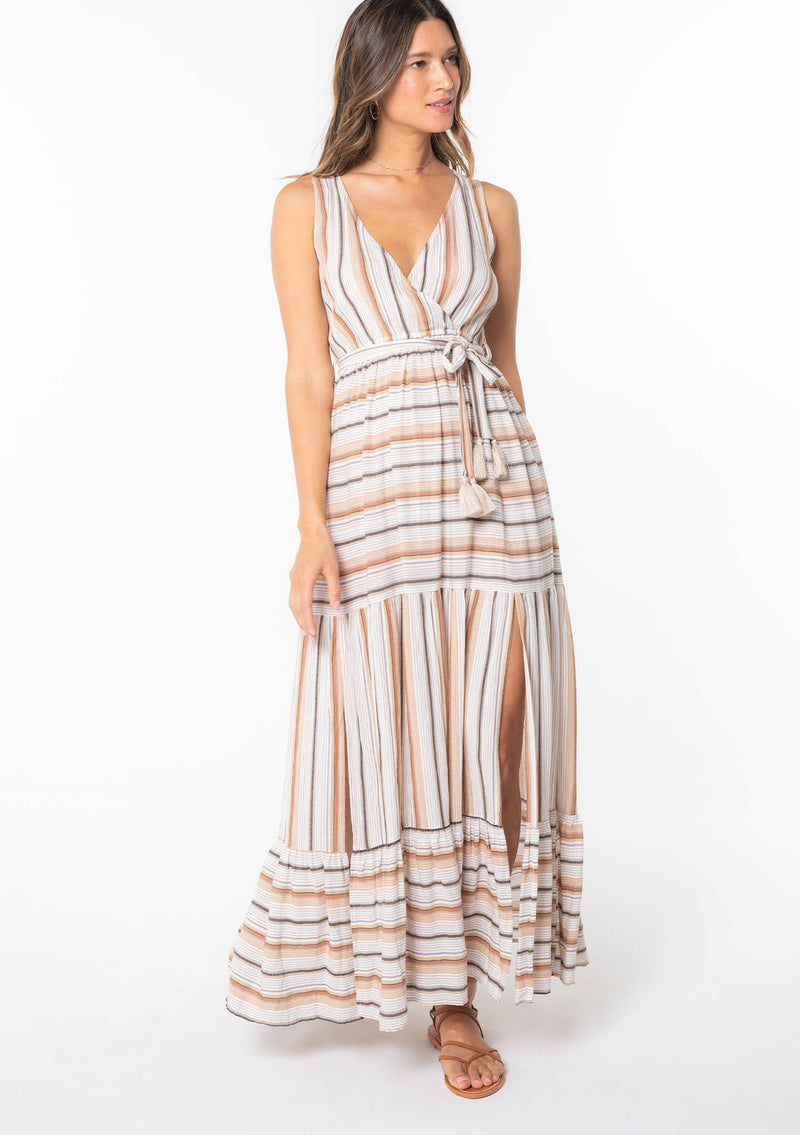[Color: Natural/Tan] A model wearing a natural and brown yarn dye stripe sleeveless maxi dress with a surplice v neckline and tiered side slit skirt. 