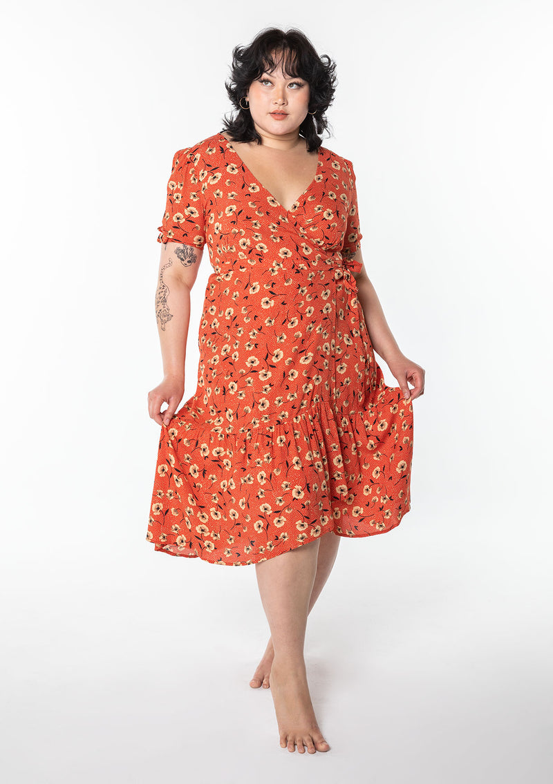 [Color: Red/Natural] A model wearing a vintage inspired mid length wrap dress in a red floral print. With short puff sleeves, a ruffled asymmetric hemline, and side tie closure.