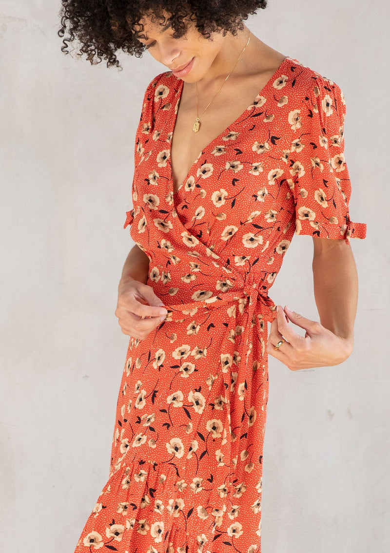 [Color: Red/Natural] A model wearing a vintage inspired mid length wrap dress in a red floral print. With short puff sleeves, a ruffled asymmetric hemline, and side tie closure. 