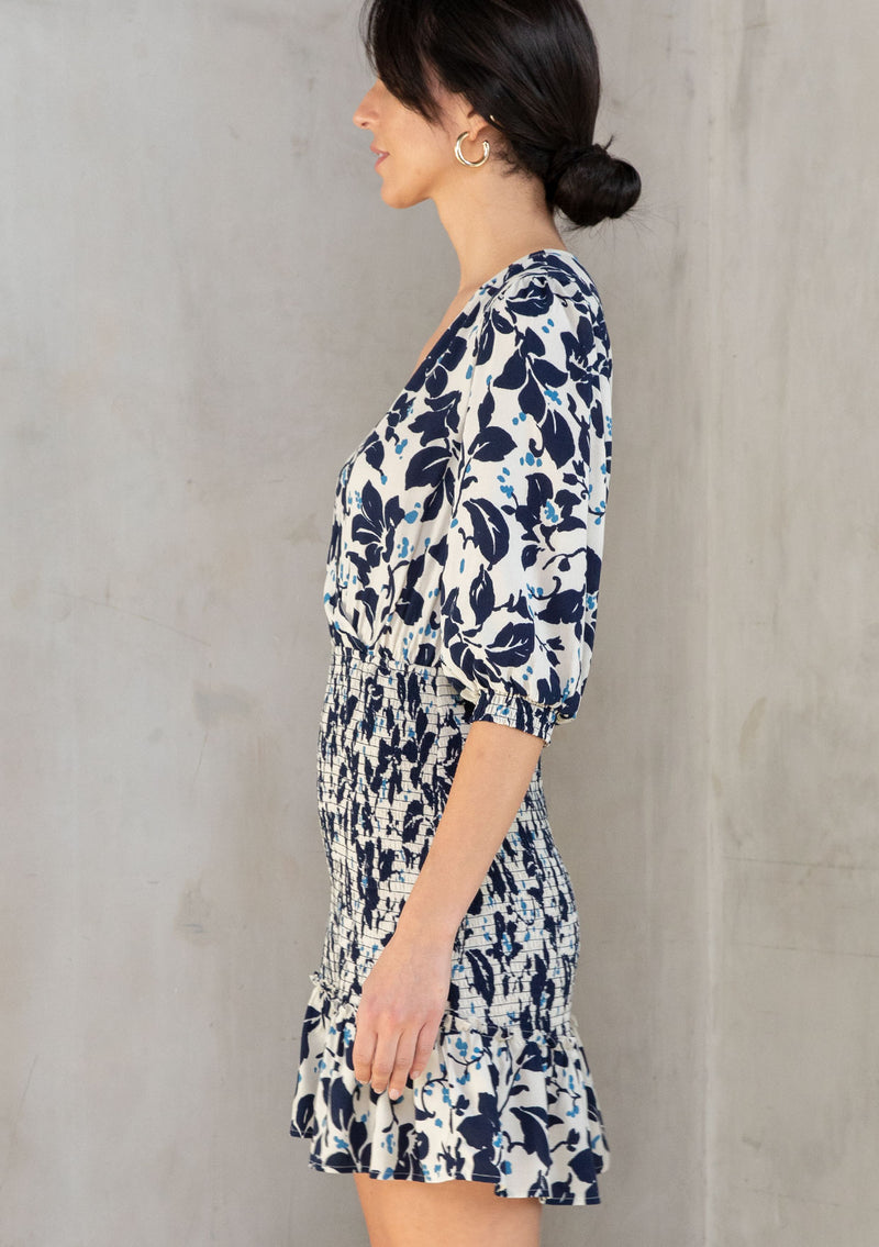 [Color: Natural/Navy] A model wearing an off white mini dress with a navy blue floral print. With a smocked skirt detail and flattering surplice v neckline. 
