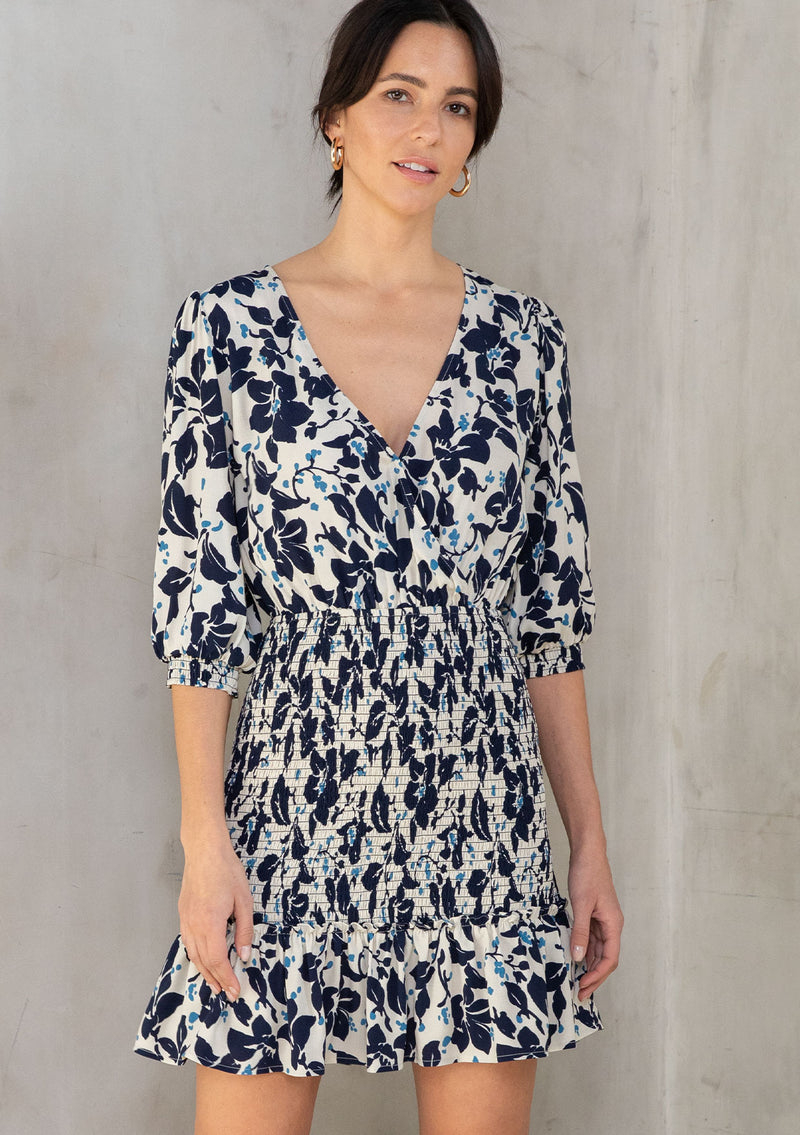 [Color: Natural/Navy] A model wearing an off white mini dress with a navy blue floral print. With a smocked skirt detail and flattering surplice v neckline. 