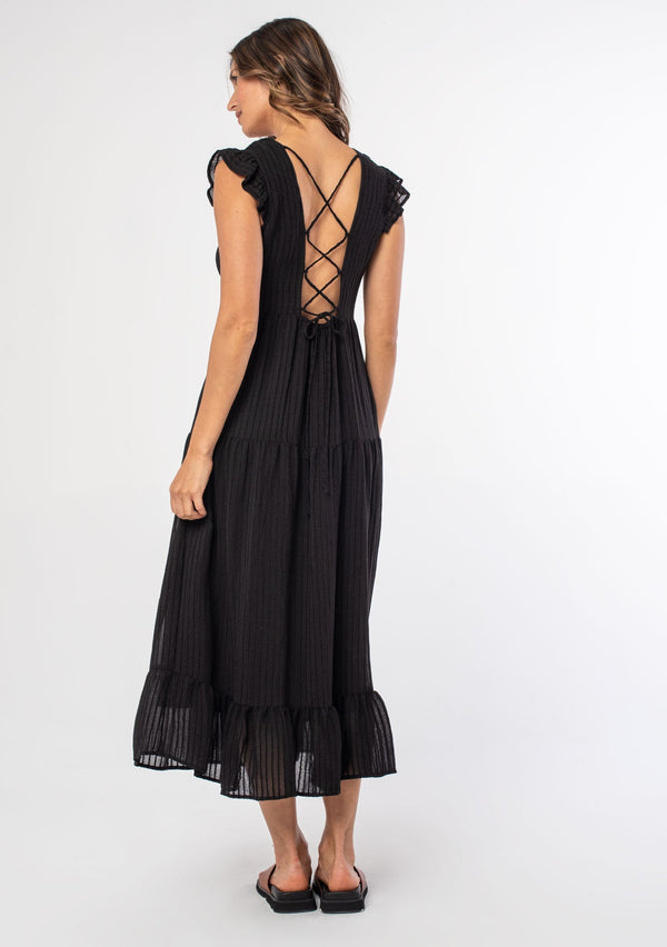 [Color: Black] A model wearing a black mid length dress with a lace up strappy back and short flutter sleeves.