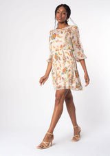 [Color: Ivory/Coral] A woman wearing a bohemian cream colored chiffon mini dress with coral floral print, a slim fit smocked skirt, and a high neckline. 