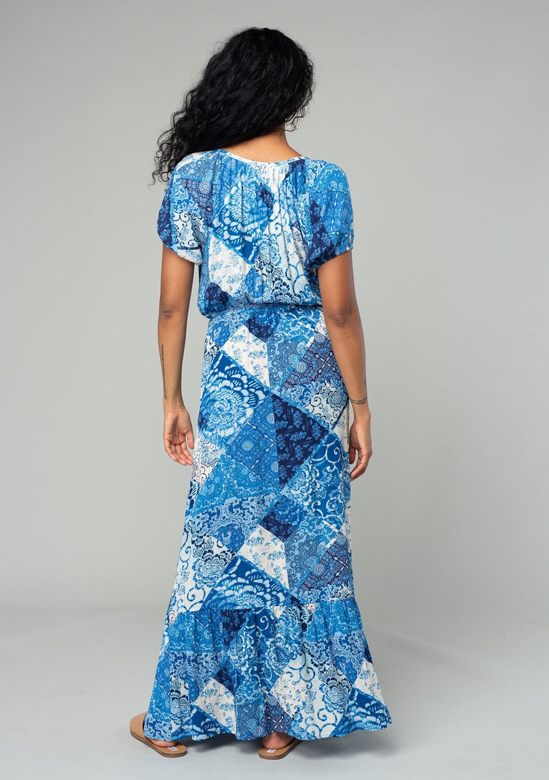 [Color: Blue/Navy] A back facing image of a brunette model wearing a bohemian flowy maxi dress in a blue and white patchwork flora print. With short puff sleeves, a v neckline, a front slit, and a tassel tie waist belt.