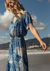 [Color: Blue/Navy] A close up side facing image of a blonde model at the beach wearing a bohemian flowy maxi dress in a blue and white patchwork flora print. With short puff sleeves, a v neckline, a front slit, and a tassel tie waist belt.