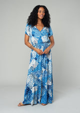 [Color: Blue/Navy] A front facing image of a brunette model wearing a bohemian flowy maxi dress in a blue and white patchwork flora print. With short puff sleeves, a v neckline, a front slit, and a tassel tie waist belt.