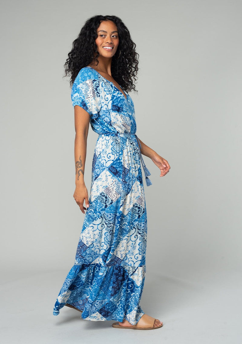 [Color: Blue/Navy] A side facing image of a brunette model wearing a bohemian flowy maxi dress in a blue and white patchwork flora print. With short puff sleeves, a v neckline, a front slit, and a tassel tie waist belt.