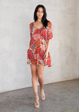 [Color: Ivory/Red] A model wearing a bohemian flowy mini dress in a red patchwork floral print. With short puff sleeves, a deep v neckline in front and back, and a tassel tie closure. 
