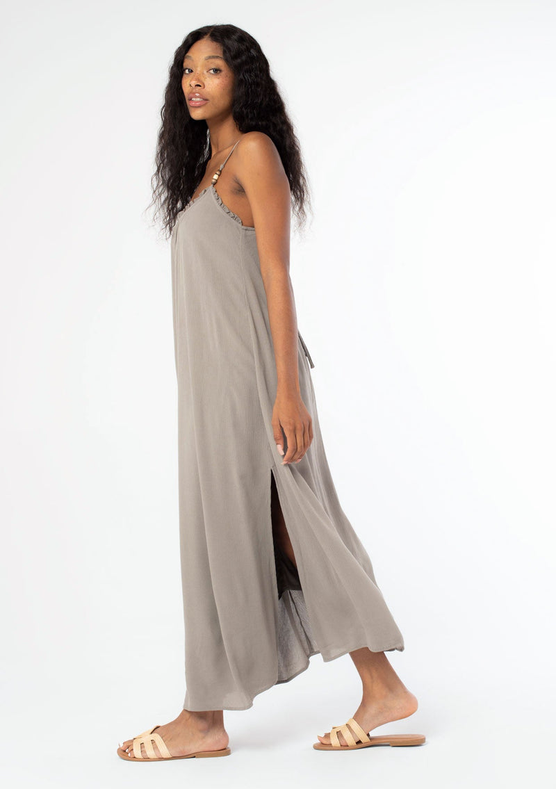[Color: Cement] A side facing image of a black model wearing a flowy grey bohemian summer maxi dress with strappy back and wooden bead details.