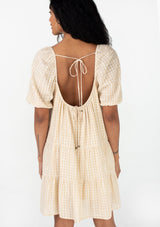 [Color: Natural] A back facing image of a brunette model wearing a cotton seersucker bohemian mini dress designed in a natural check print. With short puff sleeves, a flowy, relaxed fit, a scooped neckline, a tiered silhouette, and an open back with tie closure. 