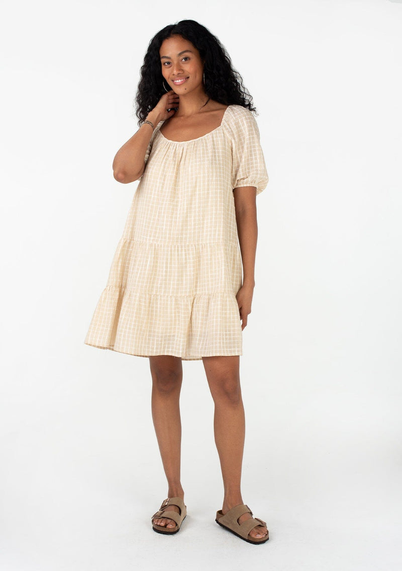 [Color: Natural] A full body front facing image of a brunette model wearing a cotton seersucker bohemian mini dress designed in a natural check print. With short puff sleeves, a flowy, relaxed fit, a scooped neckline, a tiered silhouette, and an open back with tie closure. 