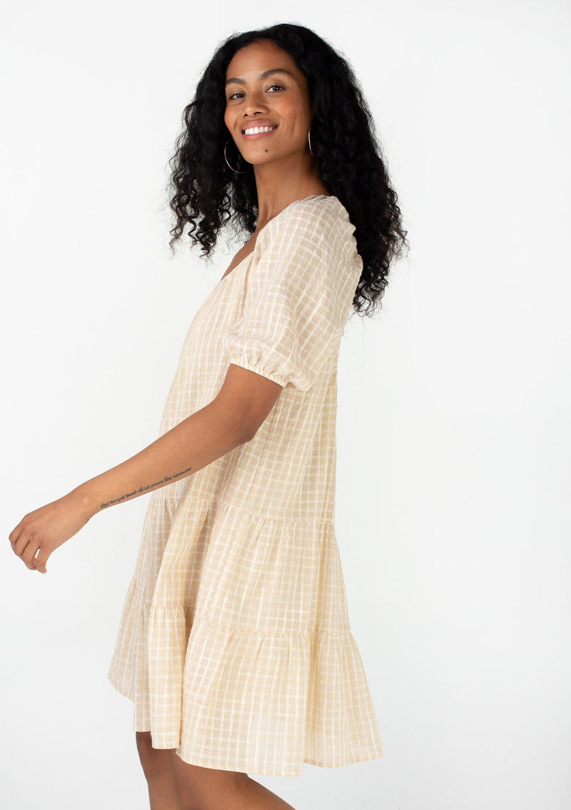 [Color: Natural] A side facing image of a brunette model wearing a cotton seersucker bohemian mini dress designed in a natural check print. With short puff sleeves, a flowy, relaxed fit, a scooped neckline, a tiered silhouette, and an open back with tie closure. 
