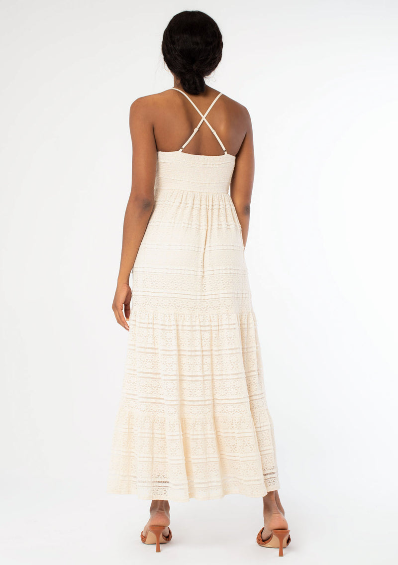[Color: Natural] A back facing image of a black model wearing a bohemian off white lace maxi dress with spaghetti straps, a flowy skirt, and a v neckline.