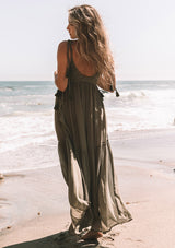 [Color: Moss Green] A back facing image of a blonde model wearing a moss green sheer maxi cover up beach dress. With adjustable tassel tie spaghetti tank top straps, a deep v neckline, a side lace up detail, and a lace trim top.