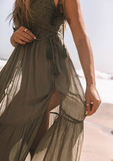 [Color: Moss Green] A close up side facing image of a blonde model wearing a moss green sheer maxi cover up beach dress. With adjustable tassel tie spaghetti tank top straps, a deep v neckline, a side lace up detail, and a lace trim top.
