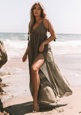 [Color: Moss Green] A front facing image of a blonde model wearing a moss green sheer maxi cover up beach dress. With adjustable tassel tie spaghetti tank top straps, a deep v neckline, a side lace up detail, and a lace trim top.