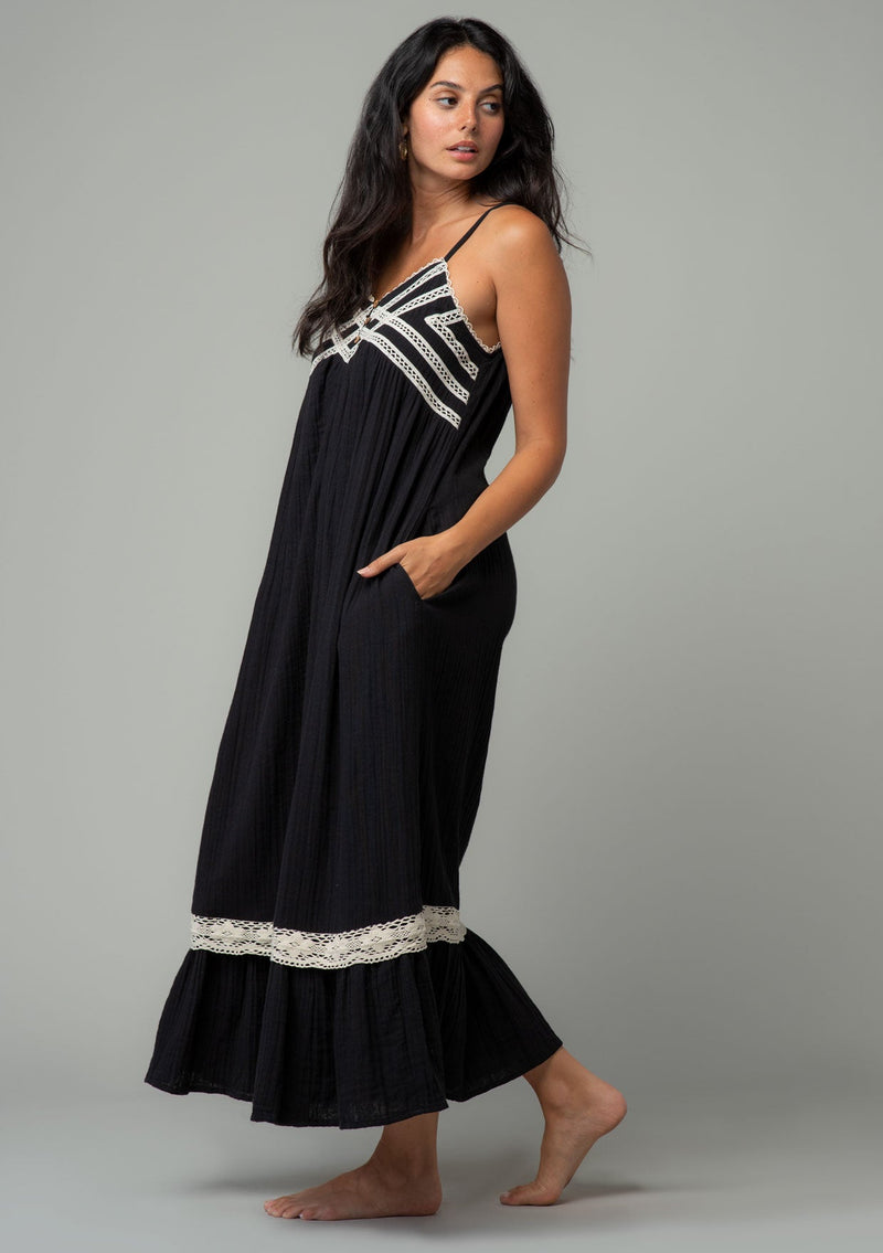[Color: Black/Natural] A full body side facing image of a brunette model wearing a black cotton gauze sleeveless maxi dress with a natural crochet trim top and hemline. With adjustable spaghetti straps and a flowy fit. 