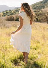 [Color: Vanilla] A back facing image of a blonde model outside in a field wearing a classic ivory bohemian spring maxi dress with eyelet lace trim throughout. Featuring short flutter cap sleeves, a v neckline, a self covered loop button front, and a tiered flowy skirt.