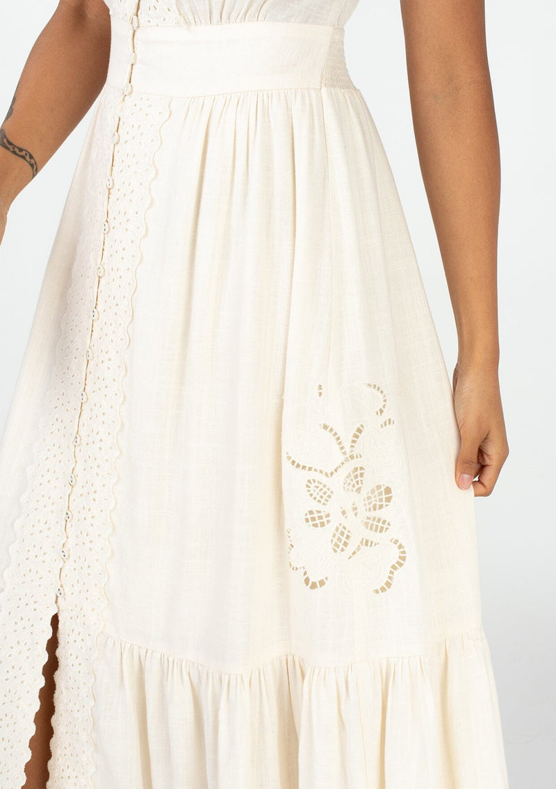 [Color: Vanilla] A close up side facing image of a brunette model wearing a classic ivory bohemian spring maxi dress with eyelet lace trim throughout. Featuring short flutter cap sleeves, a v neckline, a self covered loop button front, and a tiered flowy skirt. 