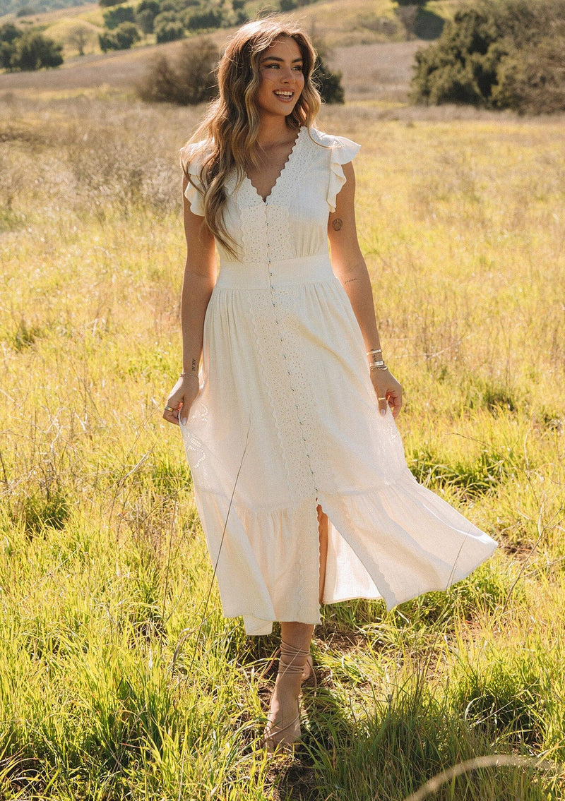 [Color: Vanilla] A front facing image of a blonde model outside in a field wearing a classic ivory bohemian spring maxi dress with eyelet lace trim throughout. Featuring short flutter cap sleeves, a v neckline, a self covered loop button front, and a tiered flowy skirt.