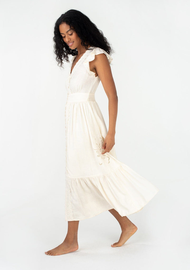 [Color: Vanilla] A side facing image of a brunette model wearing a classic ivory bohemian spring maxi dress with eyelet lace trim throughout. Featuring short flutter cap sleeves, a v neckline, a self covered loop button front, and a tiered flowy skirt. 