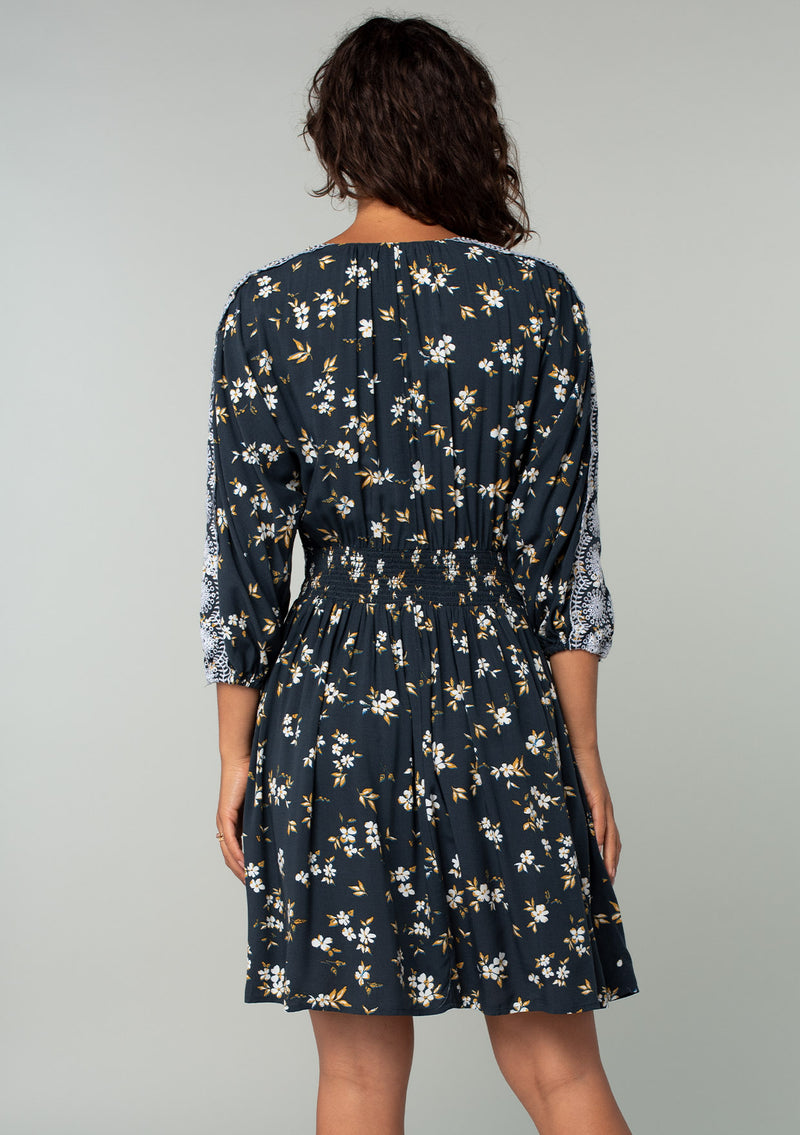 [Color: Navy/Powder Blue] A back facing image of a brunette model wearing a bohemian navy blue mini dress with a yellow and white floral print. With lace trimmed long sleeves, a button front, and a lace trimmed waist. 